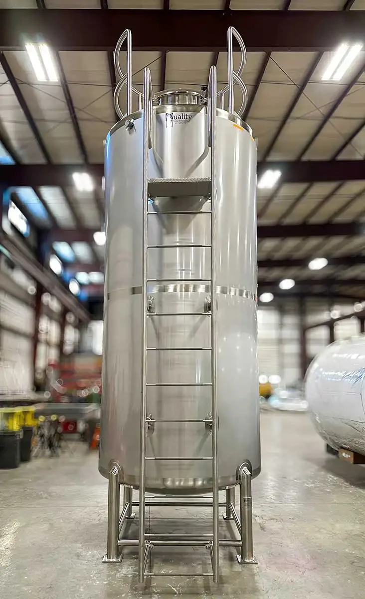 A tall cylindrical industrial storage tank with a vertical safety ladder mounted to the side. The ladder is enclosed by a protective safety cage and leads to an elevated access platform with metal guardrails at the top of the tank. The tank is constructed of stainless steel and is designed for safe access and maintenance at height.