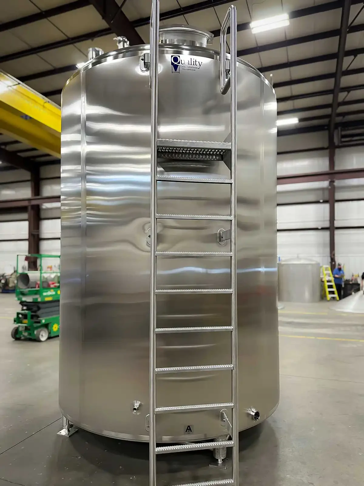 A tall cylindrical industrial storage tank with a vertical safety ladder mounted to the side. The ladder is enclosed by a protective safety cage and leads to an elevated access platform with metal guardrails at the top of the tank. The tank is constructed of stainless steel and is designed for safe access and maintenance at height.