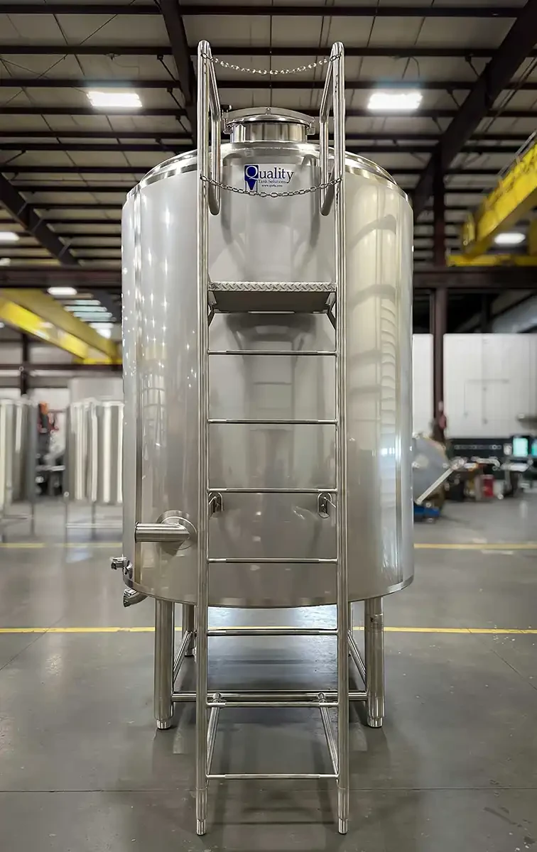 A tall cylindrical industrial storage tank with a vertical safety ladder mounted to the side. The ladder is enclosed by a protective safety cage and leads to an elevated access platform with metal guardrails at the top of the tank. The tank is constructed of stainless steel and is designed for safe access and maintenance at height.